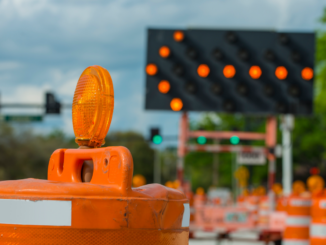Photo shows a left arrow message board in the background and orange construction barrels in the foreground (stock photo).